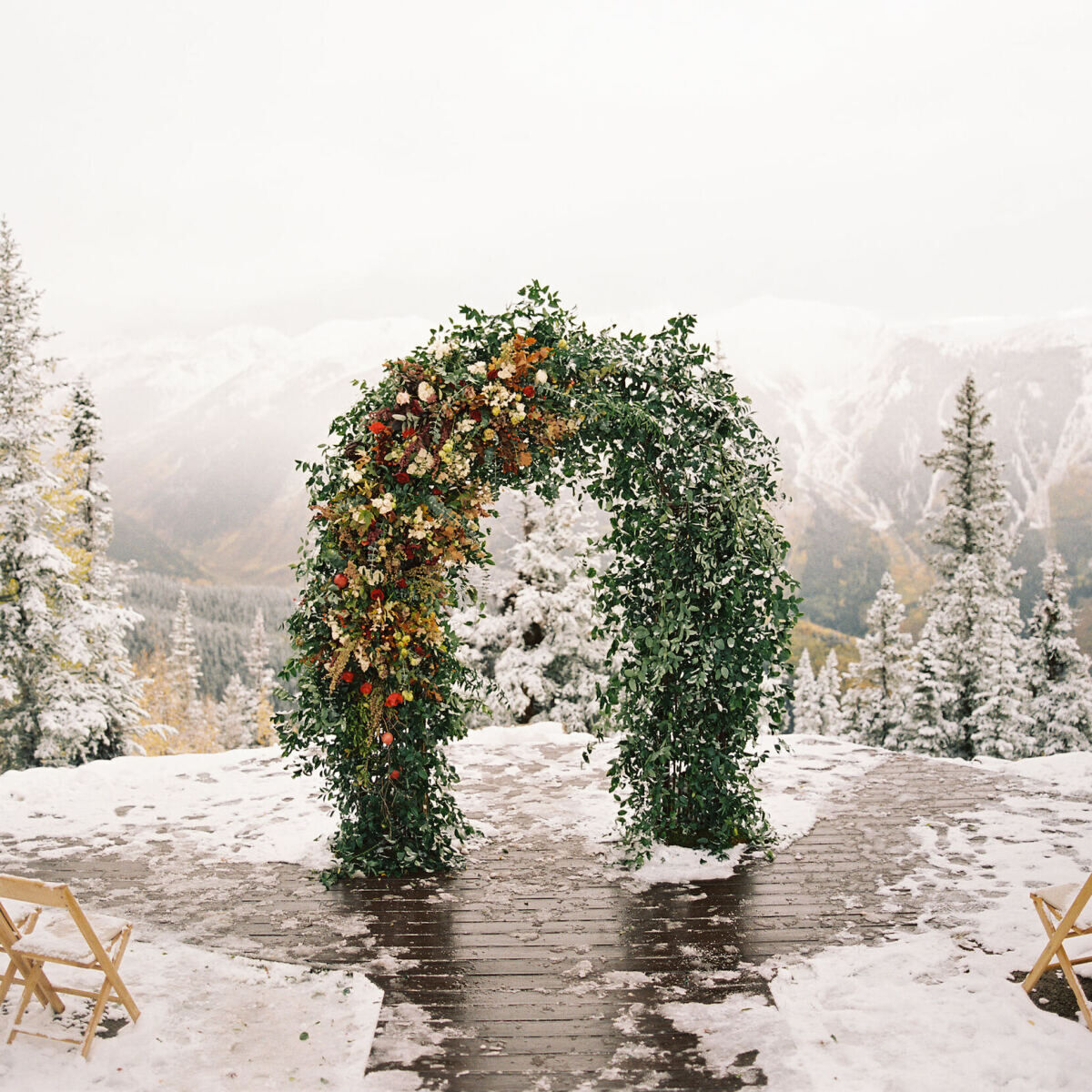 Holiday Wedding: An outdoor snowy wedding ceremony set-up with pine trees in the background. There's a wedding arch adorned with greenery and red florals.