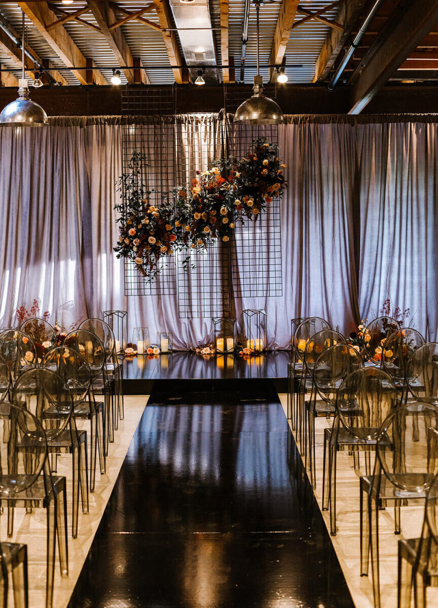 An industrial wedding ceremony, with a floral installation attached to a grid and smoke grey ghost chairs lining the mirrored aisle.