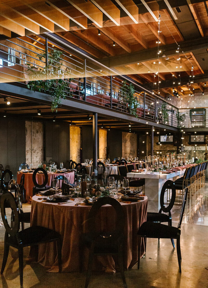 An industrial wedding venue, set for a reception, complete with greenery hanging from the balcony, strands of bistro bulbs to illuminate the space, and a mix of table shapes and sleek black chairs.