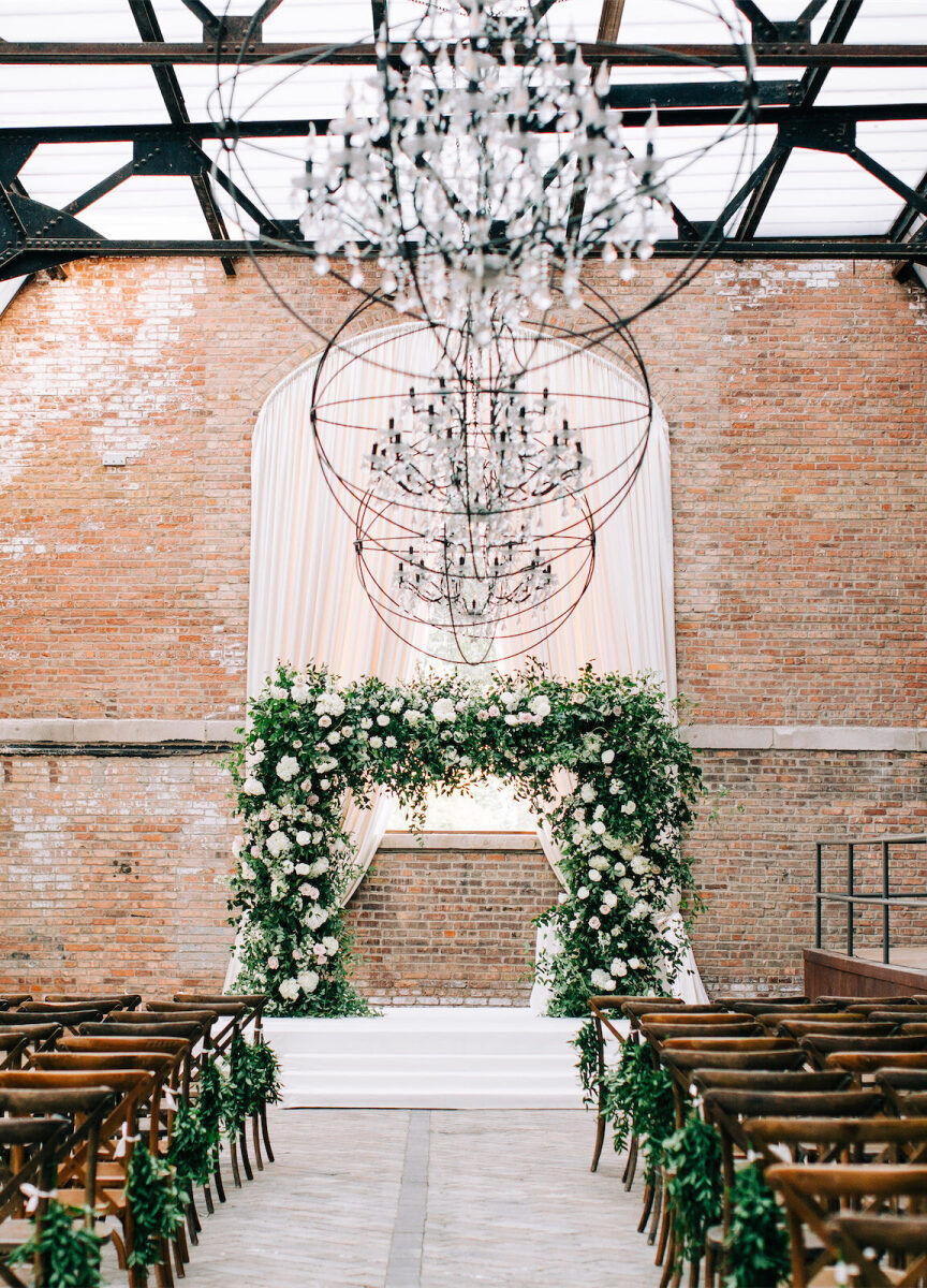 Industrial Wedding Venues: A ceremony set-up with a greenery archway against a brick backdrop. There are three unique, industrial light fixtures hanging form the ceilings and wooden chairs for guests.