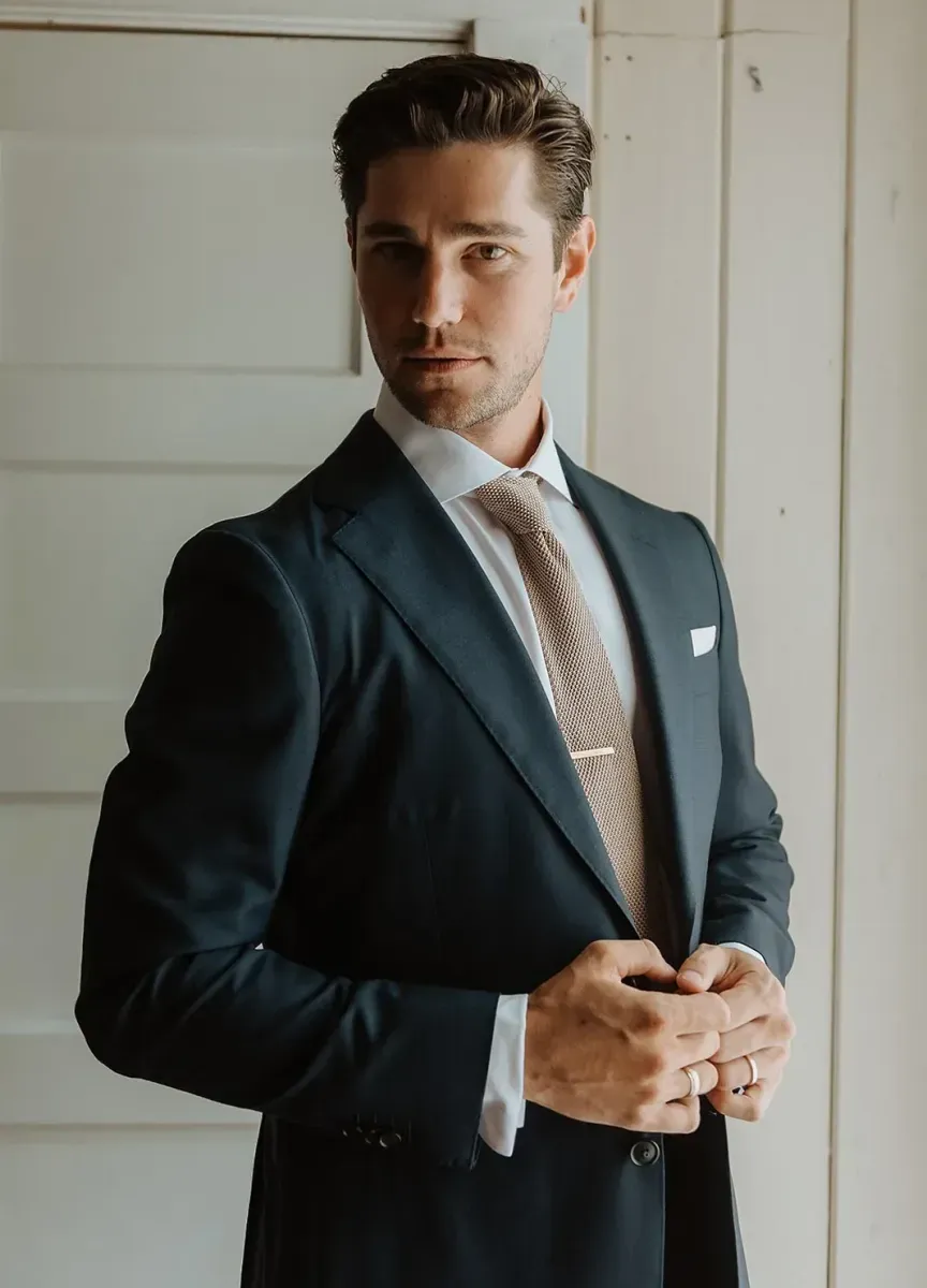 A groom looking directly at the camera and holding his suit dark suit jacket, with a button down shirt and neutral-toned tie. 