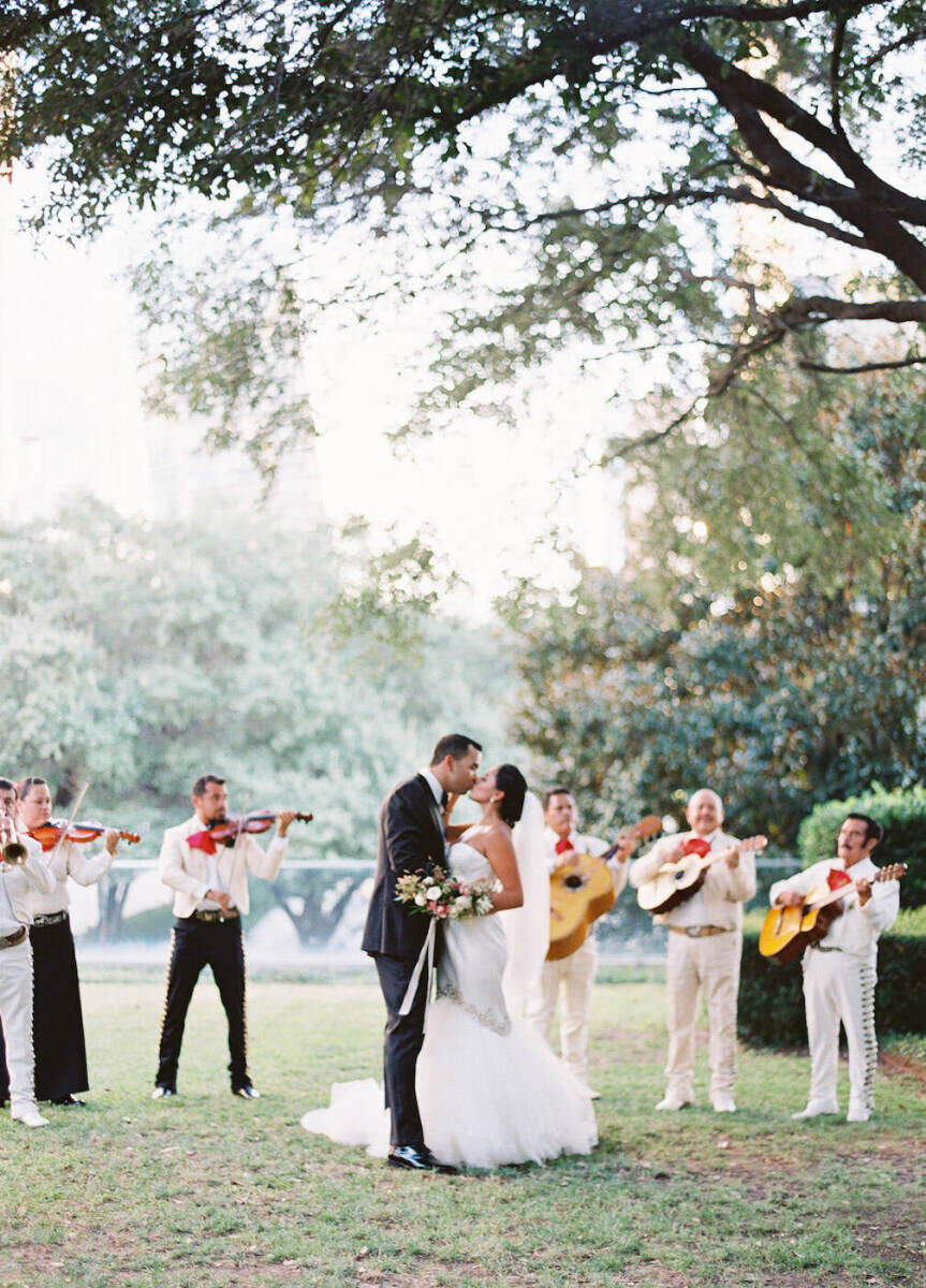 Latinx Weddings: A bride and groom kissing outdoors with a mariachi band playing behind them.