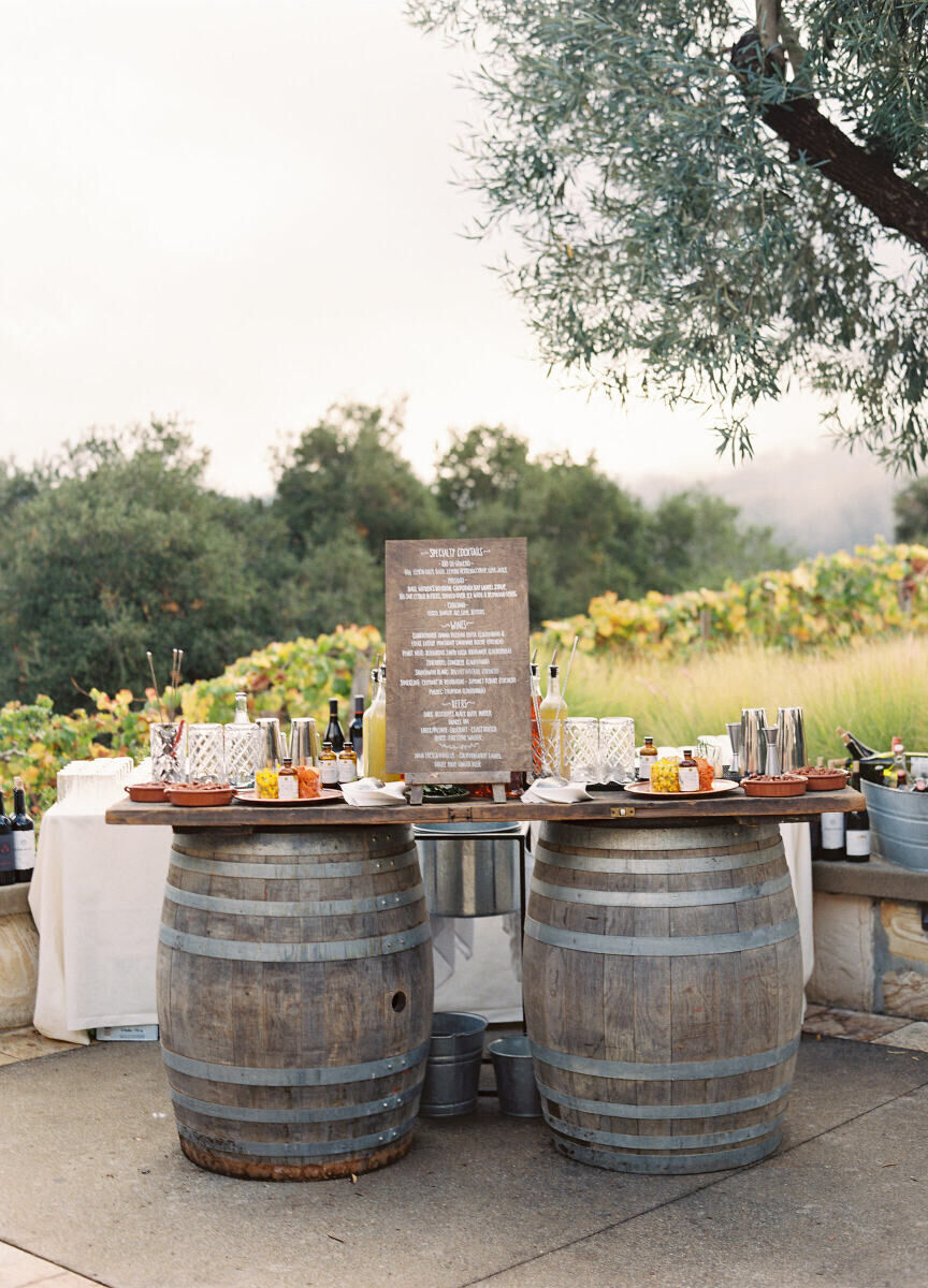 Latinx Heritage: A bar setup with two barrels and a sign with the drink menu at an outdoor reception.