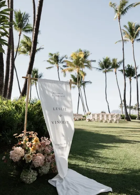 A welcome sign billowing in the wind with flowers on the ground and an outdoor wedding ceremony setup and palm trees in the distance at a wedding venue in Hawaii. 