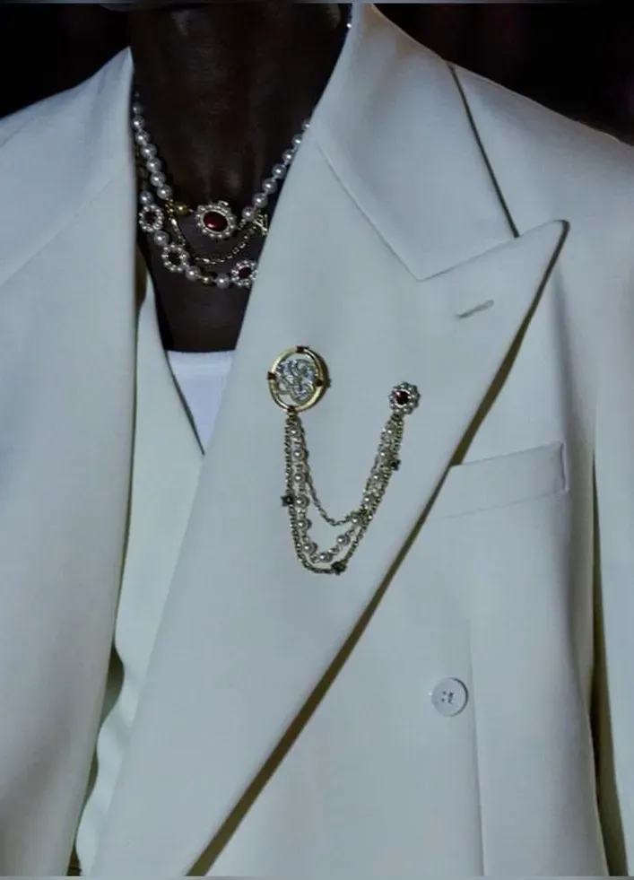 White suit with a single jeweled lapel brooch and a draped crystal chain over layered necklaces. 