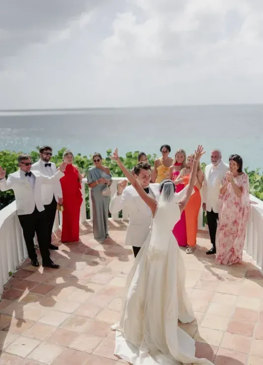 A wedding couple celebrating with their hands up and guests cheering all around them at a Caribbean wedding venue overlooking the ocean.