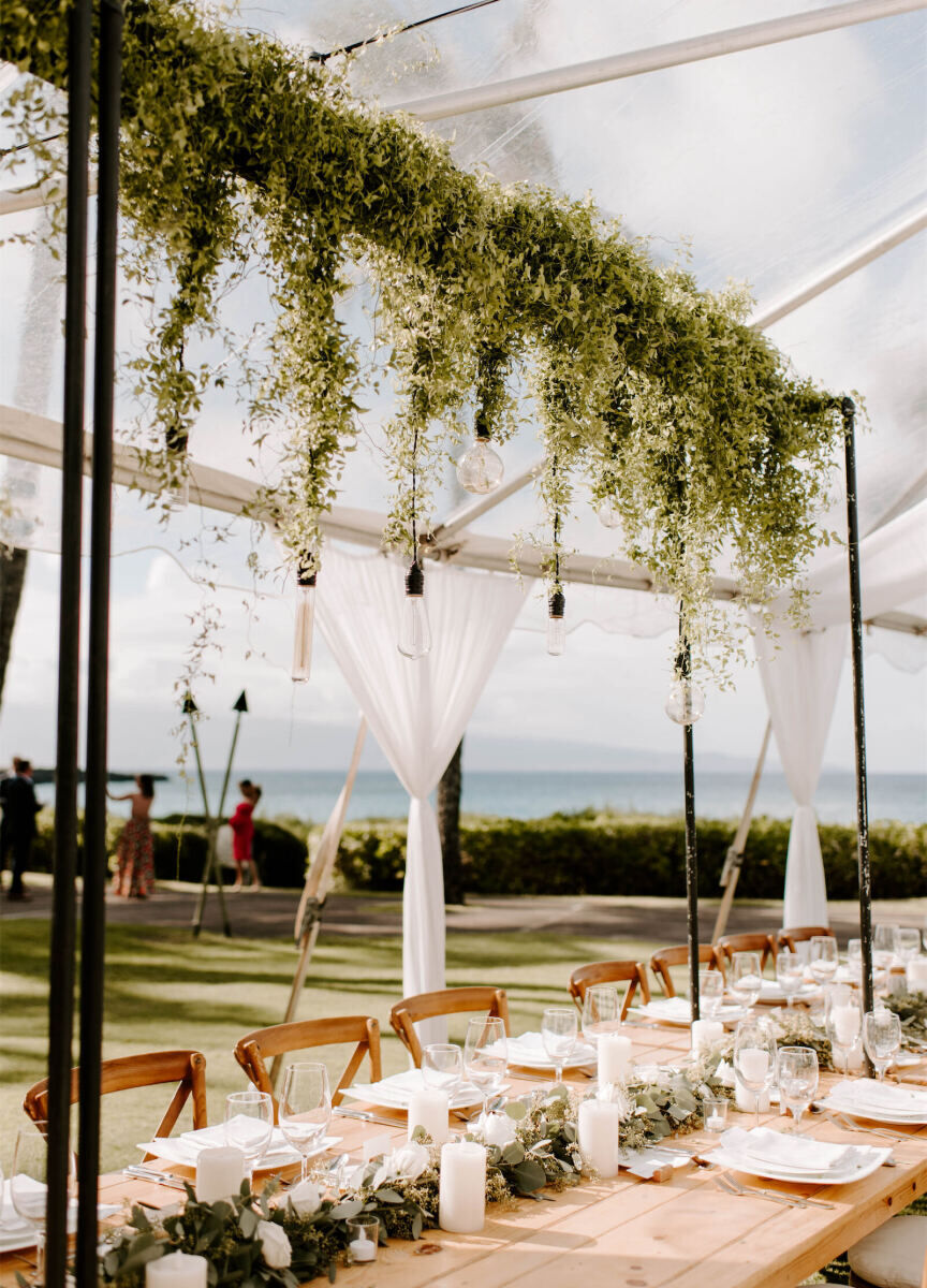 Marriott Weddings: An outdoor reception setup with greenery hanging from the ceilings of a clear tented structure overlooking the ocean in Hawaii.