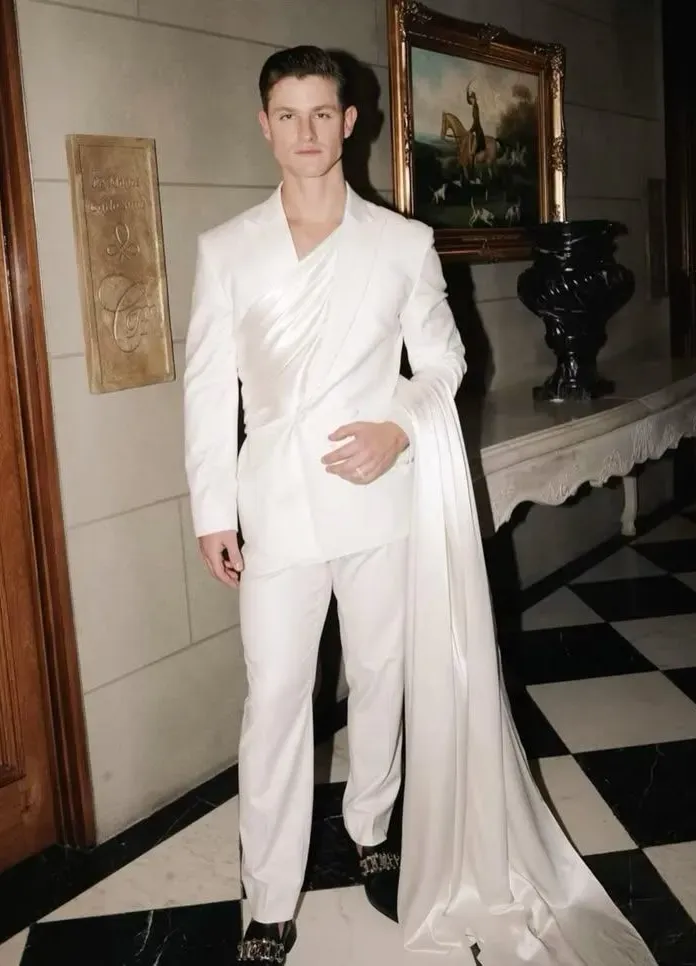 Full-length white suit with a draped satin sash, photographed in a checkerboard-floor interior. 
