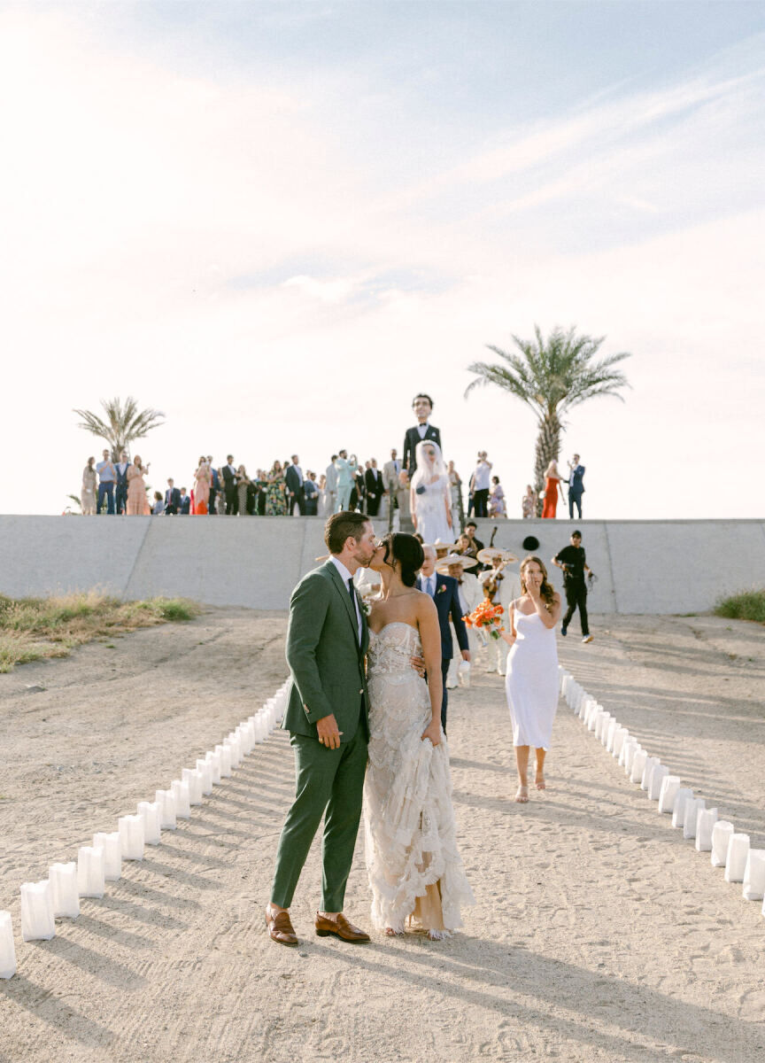 Mexican wedding: couple kissing on a dirt path followed by  their Mojiangas and wedding guests 