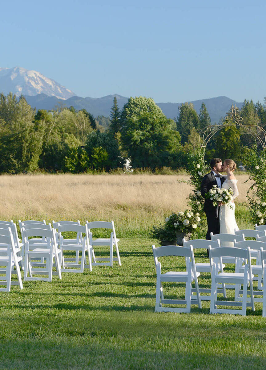 Mountain Wedding: A wedding couple kissing at a ceremony setup with white chairs, and trees and a snow-capped mountain in the background.