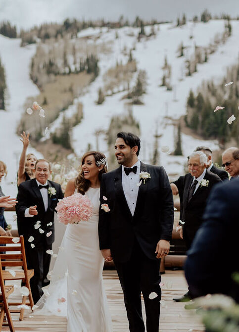 Mountain: A wedding couple walking back up the aisle after their outdoor ceremony with a view of a snow-covered mountain behind them.