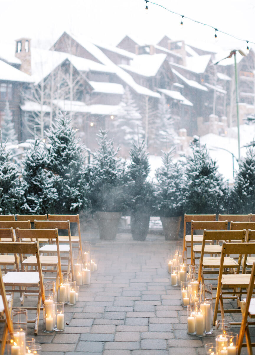 Mountain Wedding: An outdoor wedding ceremony setup with wooden folding chairs, candles as the aisle decor and snow-capped trees and buildings in the background.