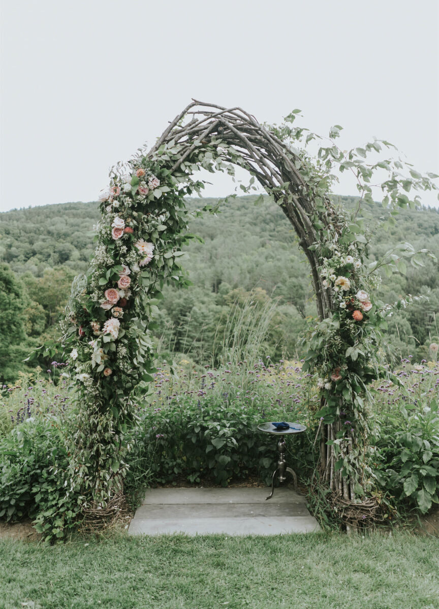 Mountain Wedding: A natural, wooden arch covered in greenery and flowers with a small table nearby and mountains in the background.