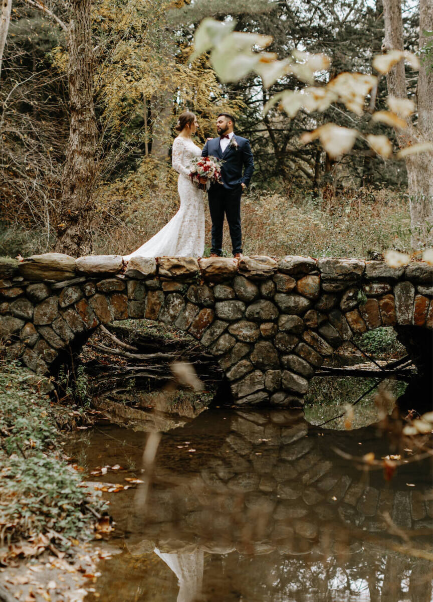 Mountain Wedding: A wedding couple smiling at each other while standing on a stone bridge over a body of water surrounded by trees.