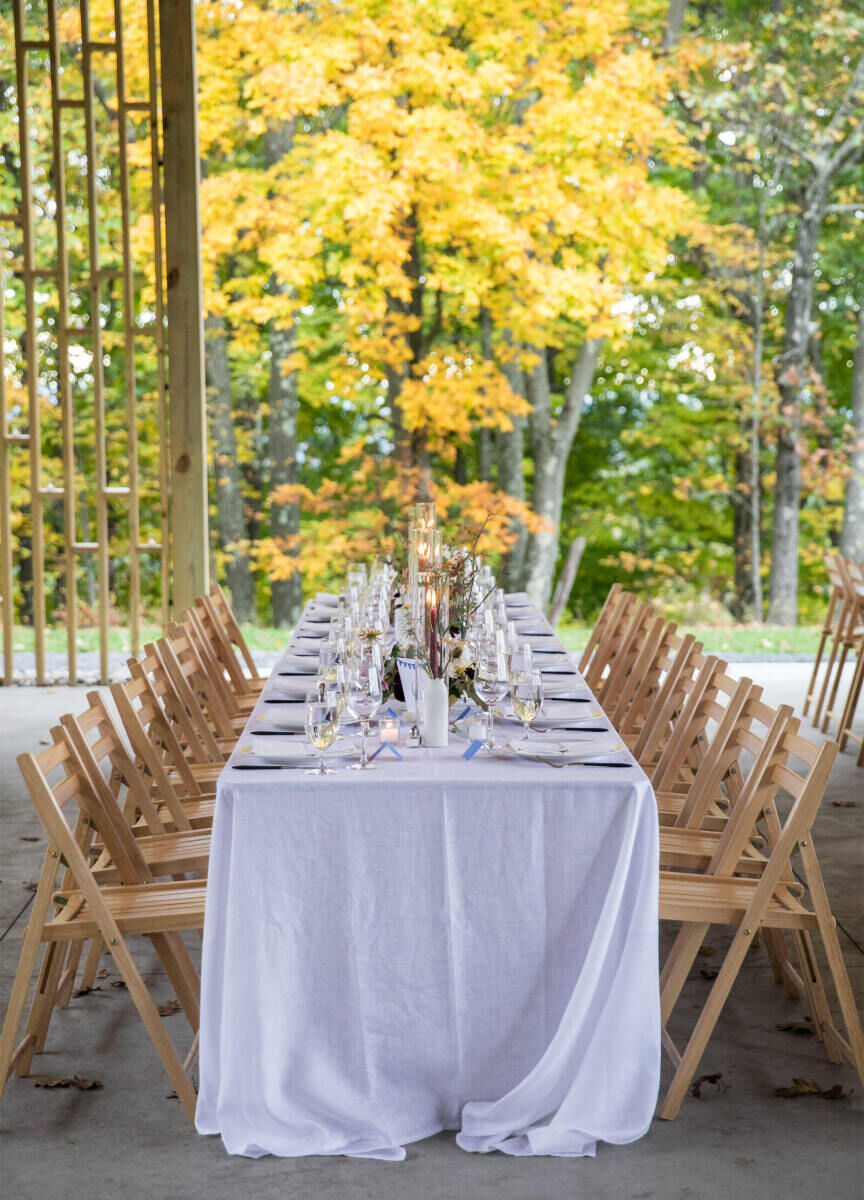 Mountain Wedding: An open indoor reception area with a long rectangular table with a white tablecloths and wooden chairs, and a view of the trees outside.