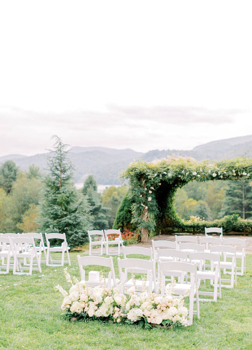 Mountain Wedding: An outdoor ceremony setup with white chairs, florals, and an oversize altar with greenery in the shape of an arch and mountains in the background.