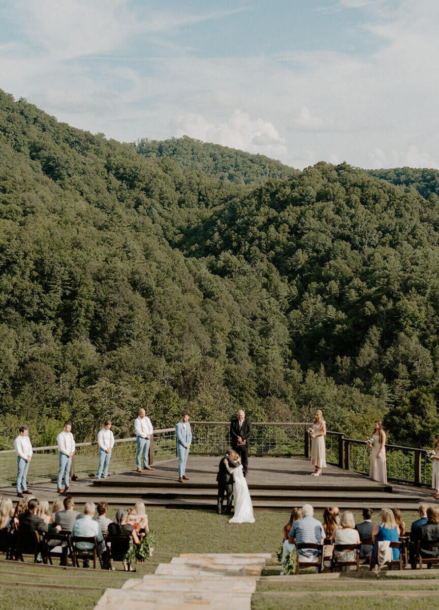 Mountain Wedding: A faraway shot of a mountainside outdoor wedding ceremony with several rows of guests, an officiant and wedding party at the altar, and everyone is surrounded by tree-covered mountains.