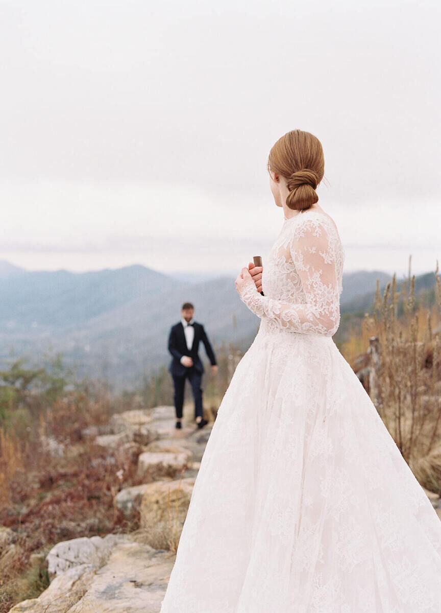 Mountain Wedding: A bride in a long-sleeve bridal gown in the foreground, with a groom walking toward her on a rocky pathway and mountains in the background.