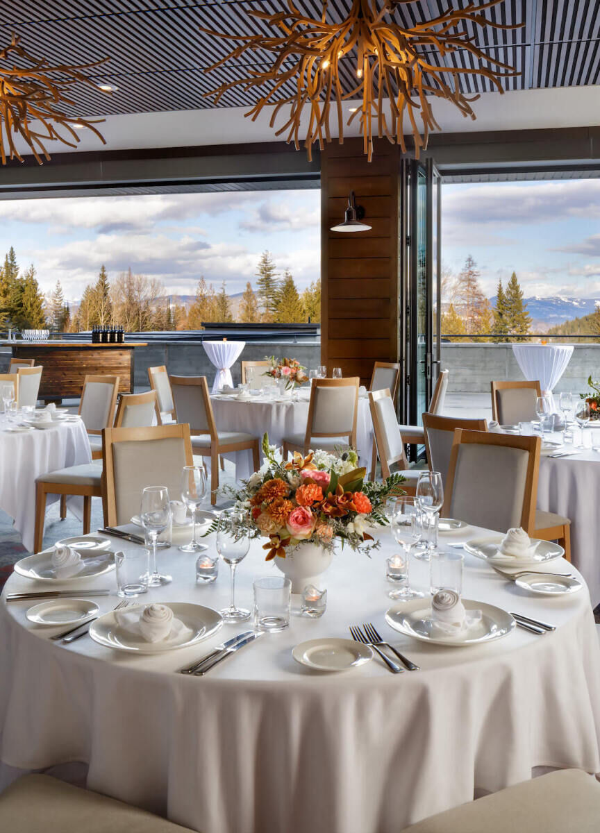 Mountain Wedding: An indoor reception setup with round tables and eclectic chandeliers and views of trees and mountains outside.