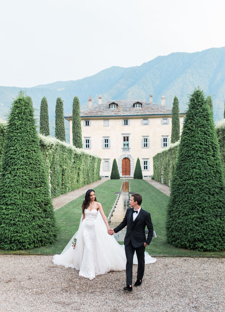 Mountain Wedding: A wedding couple holding hands and smiling in between two hedged trees, at the top of a lawn leading up to an estate in the mountains of Lake Como.