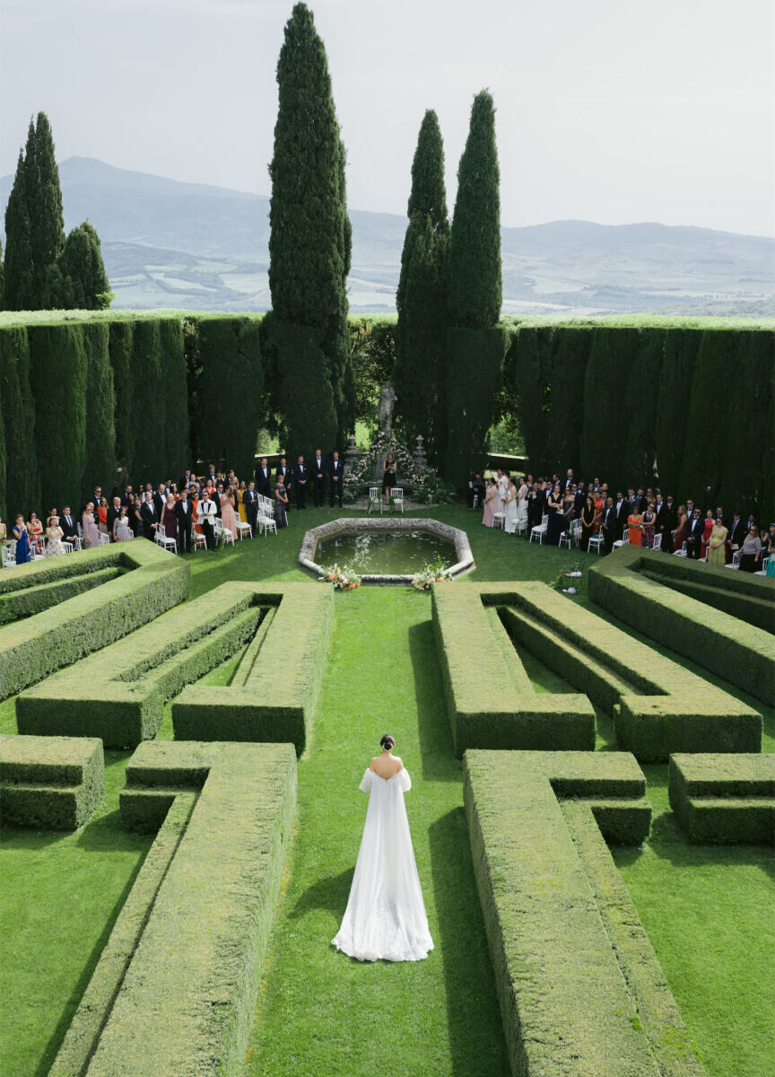 Mountain Wedding: A bride walking down a manicured garden pathway, creating by hedging, leading up to a small pond and altar just beyond it, with mountains and trees in the background.