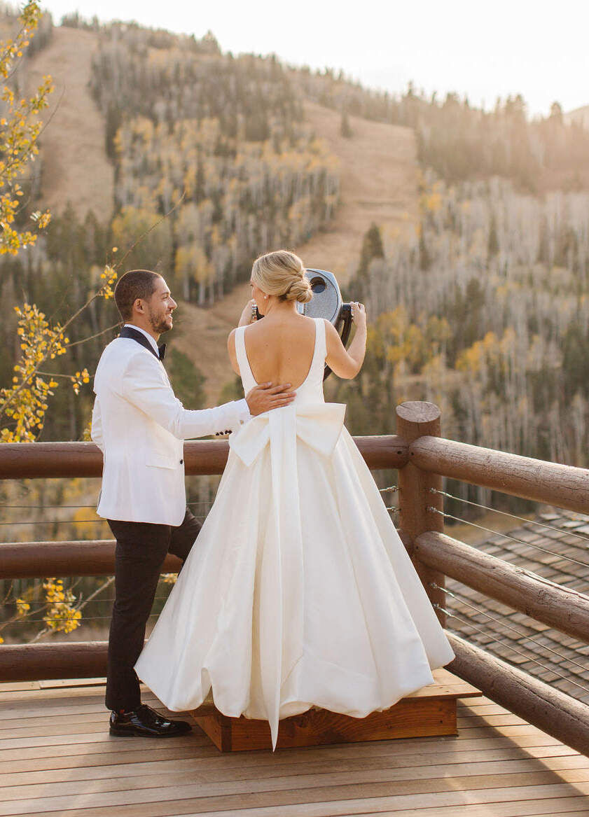 Mountain Wedding: A groom with his arm on a bride's back as she prepares to look through a telescope in the mountains.