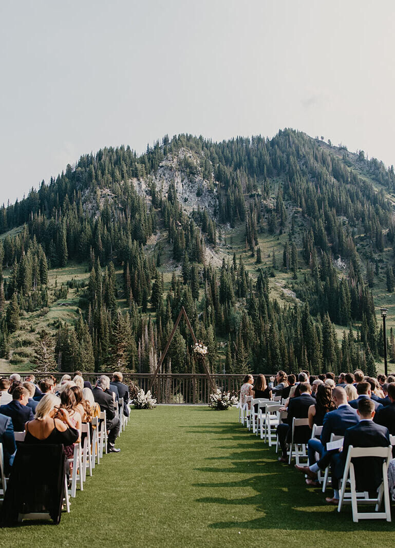 Mountain Wedding: An outdoor reception setup on a grass lawn with guests seated in white chairs and a mountain with trees all over it in the background.