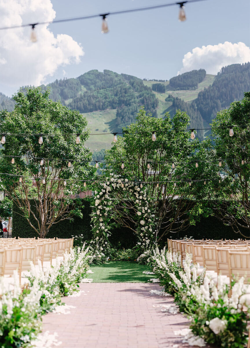 Mountain Wedding: An outdoor wedding ceremony setup with gold chairs and low-to-the-ground white floral arrangements lining the aisle, with an arch at the end, string lights above, and a green mountain in the background.