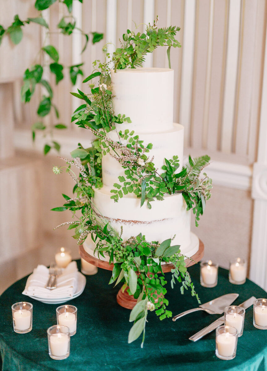 Mountain wedding details: A semi-naked wedding cake covered with fresh greenery, on display atop a green velvet linen decorated with votive candles.