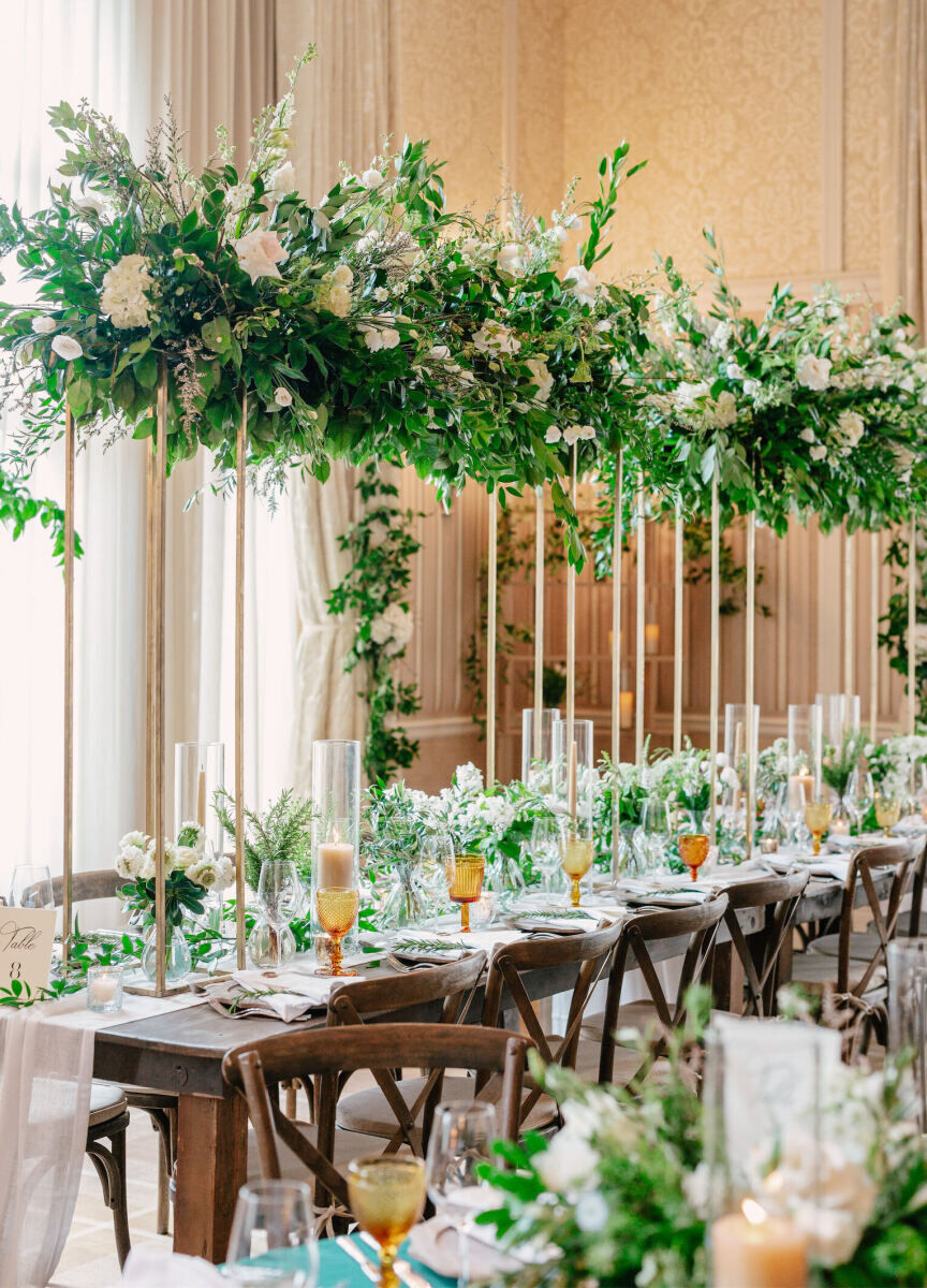 Mountain wedding details: Tall floral arrangements soar over farm tables set with a gauzy table runner, amber glassware, and pillar candles at a ballroom wedding reception.