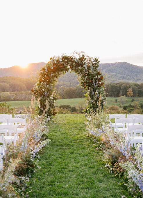 Pippin Hill Farm: A ceremony setup with white chairs, floral arrangements lining the aisle and an arch with greenery and some flowers.