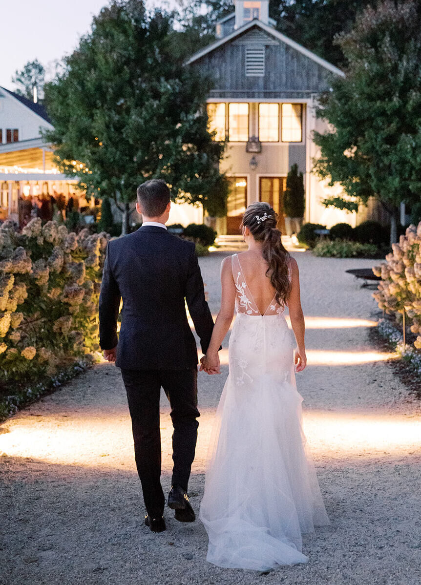 Pippin Hill Farm: A wedding couple holding hands while walking along a hydrangea-lined walkway, toward a barn.