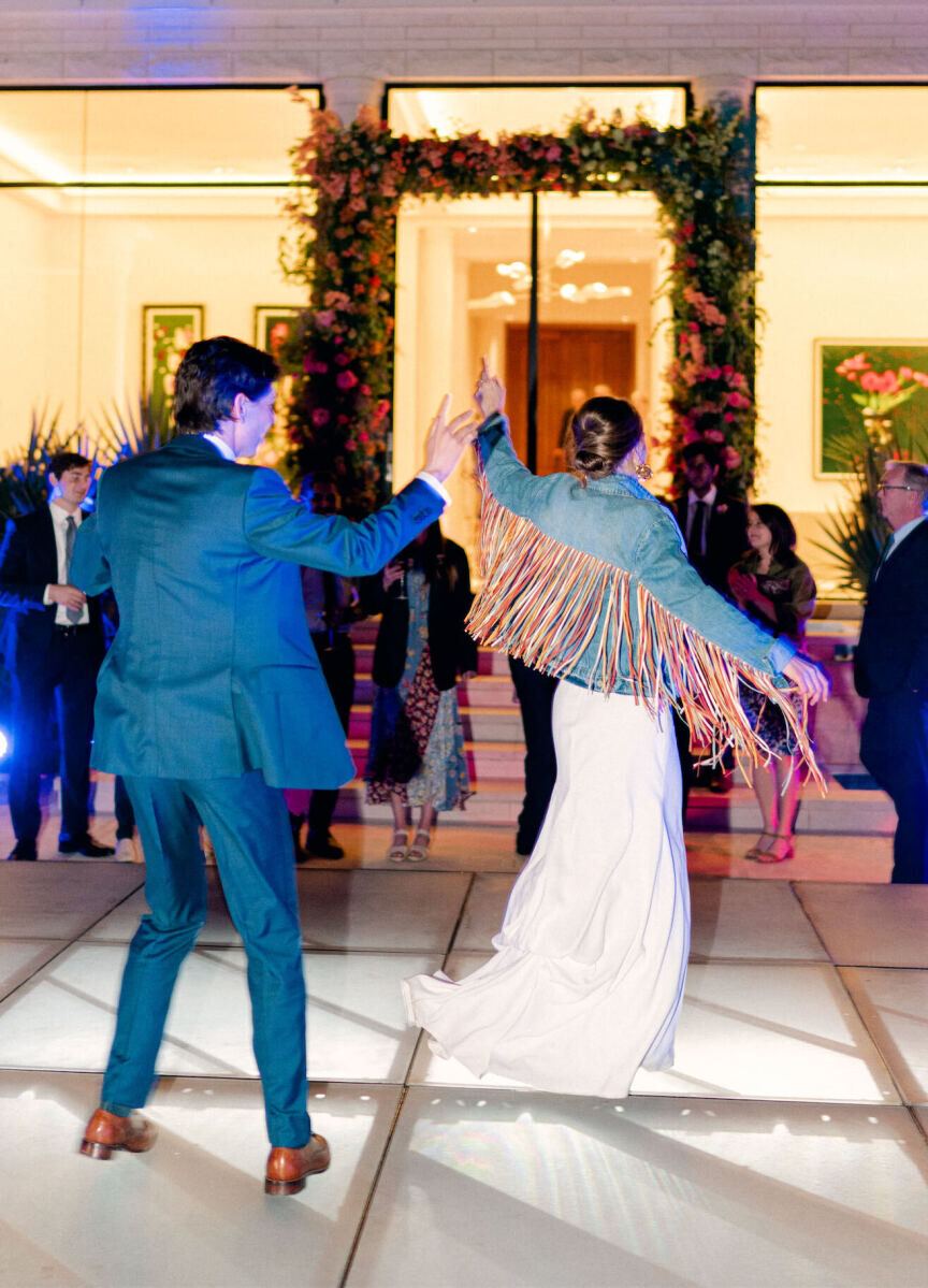 A groom spins his bride (wearing a fringed jean jacket) on the dancefloor during their rainbow wedding reception.