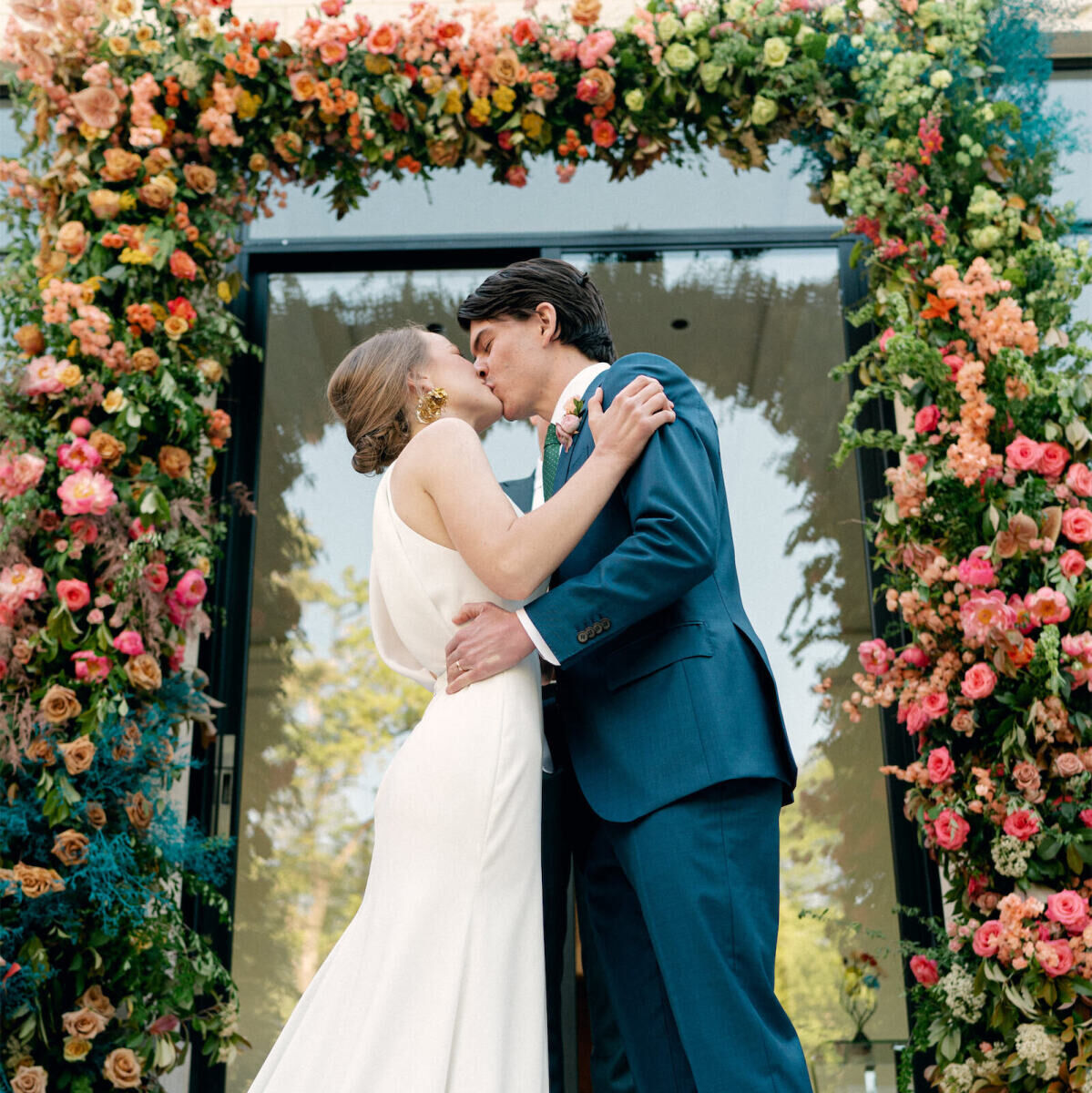 A bride and groom kiss under their rainbow wedding ceremony arch.