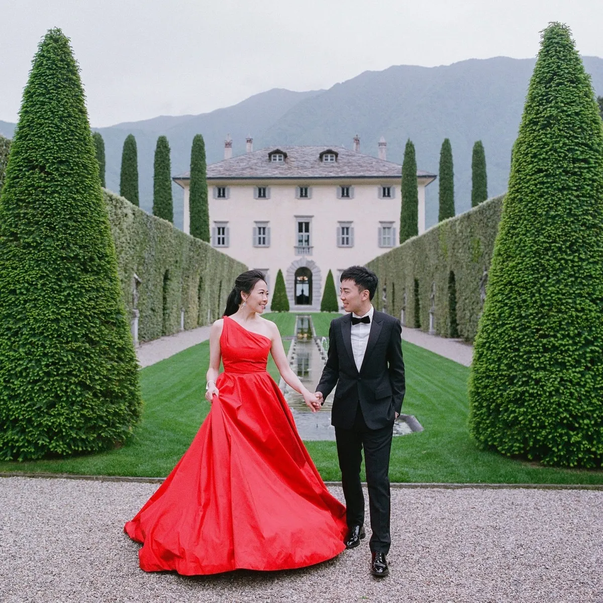 Bride in red wedding dress, wedding couple holding hands in front of an estate. 