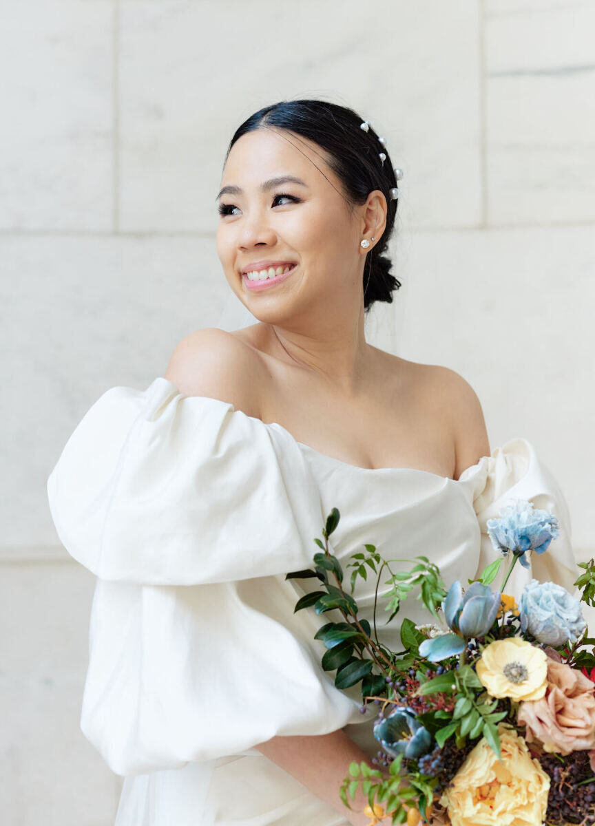 A smiling bride, in an off-the-shoulder, puff-sleeve wedding dress and pearl hair pins in her sleek and low updo.