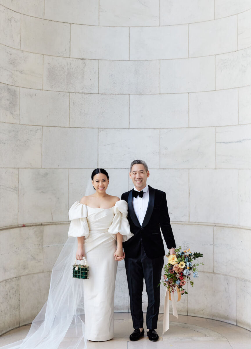 A bride wearing and off-the-shoulder dress with puff sleeves and holding a green purse, and a groom in a tuxedo and holding her colorful bouquet, pose for a portrait in front of the New York Public Library before heading to their restaurant wedding reception at Eleven Madison Park.