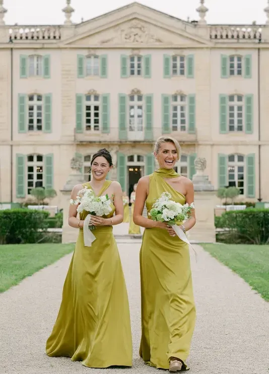 Two bridesmaids walking down an outdoor pathway wearing green gowns and holding green and white floral bouquets at a chateau wedding in France.