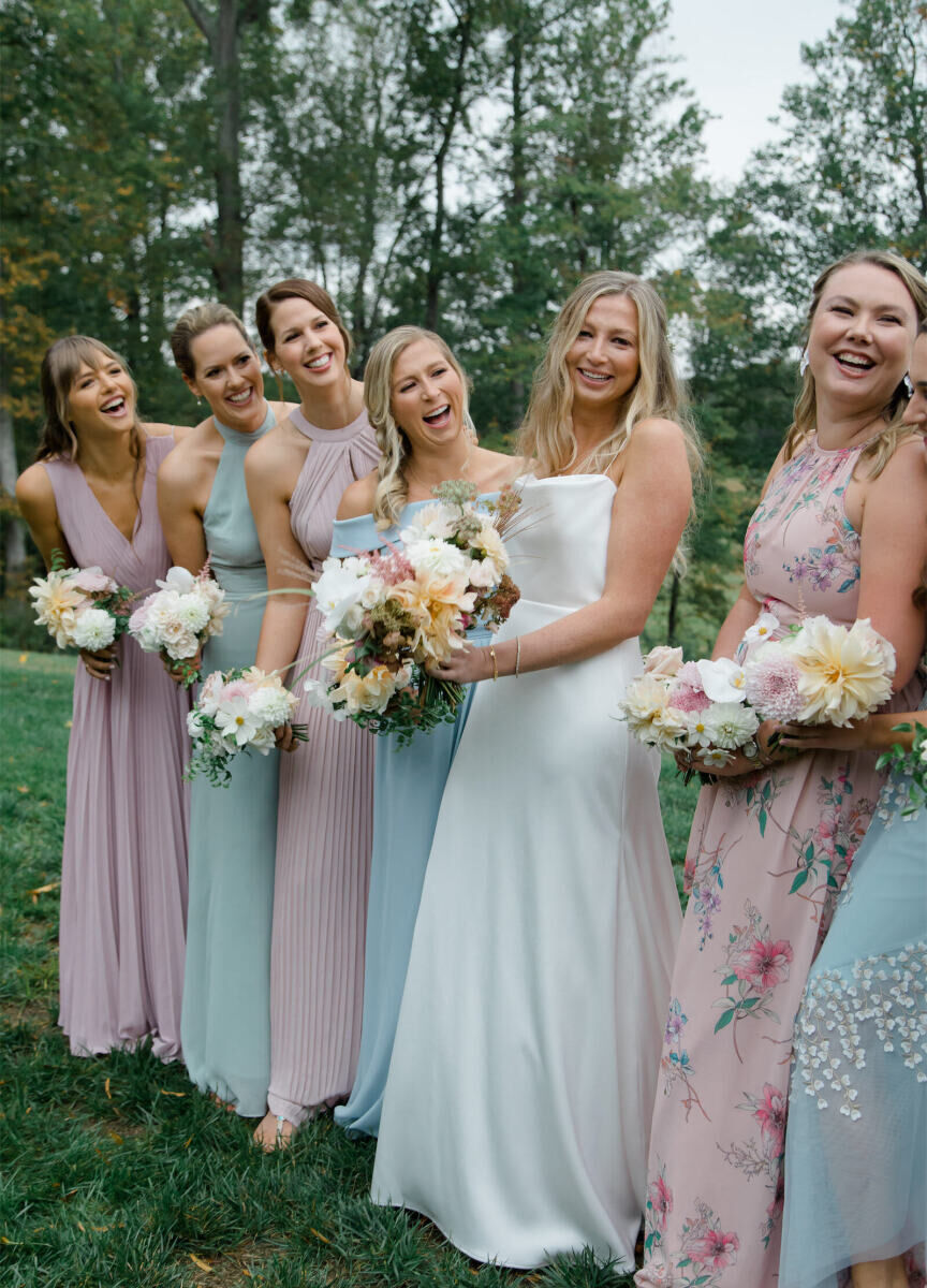 A bride and her bridesmaids—wearing mismatched pastel dresses—share a laugh during pre-ceremony portraits at her rustic outdoor wedding.