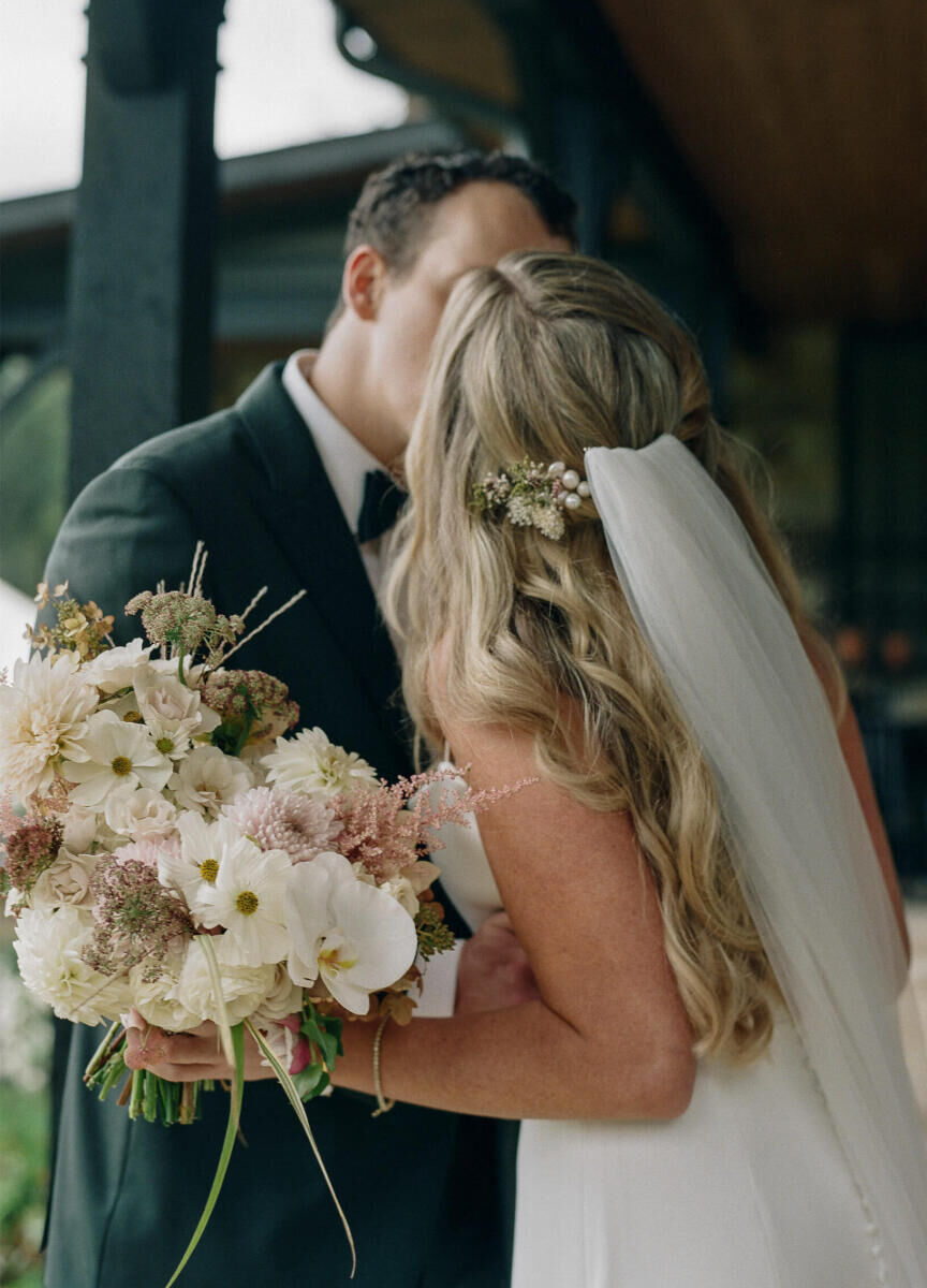 A groom and bride (wearing her hair half up and adorned with fresh flowers and pearl hair pins) kiss on the day of their rustic outdoor wedding.