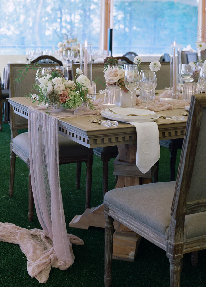 An elegant reception table at a rustic outdoor wedding was set with monogrammed dinner napkins, upholstered chairs, and soft pink gauzy table runners.