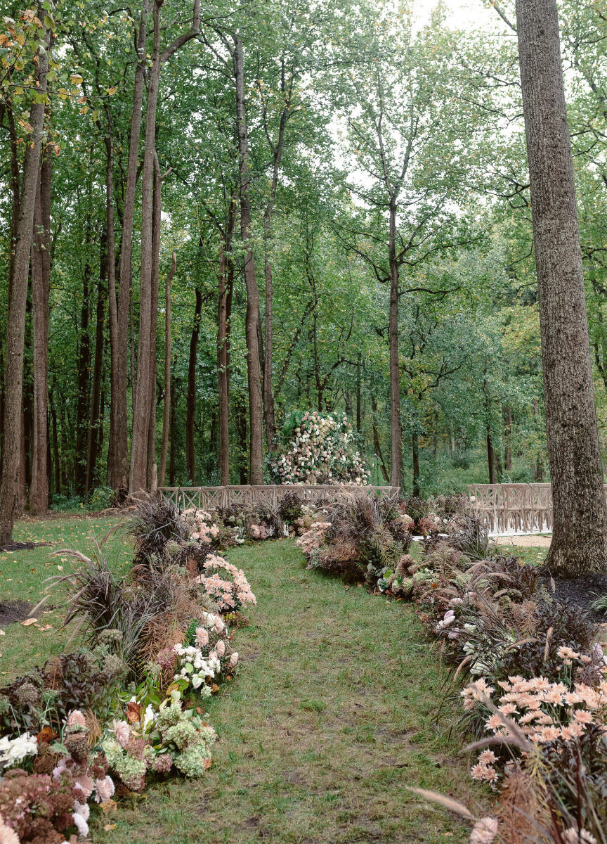 A curved aisle leading to the ceremony of a rustic outdoor wedding in the woods was decorated with flowers in soft pink, muted green, and neutrals like brown and white.