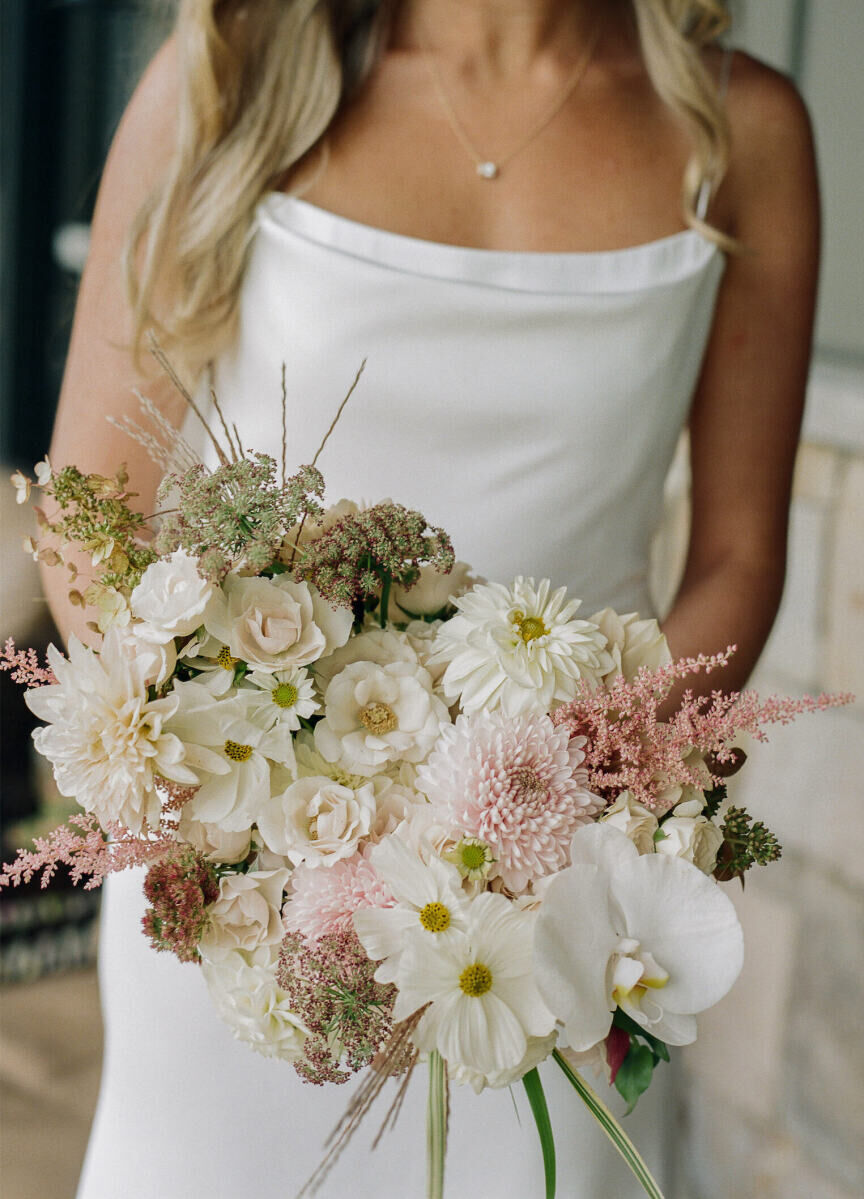 At her rustic outdoor wedding, a bride holds her white and pink bouquet, which included a mix of orchids, astilbe, dahlias, ranunculus, and roses.