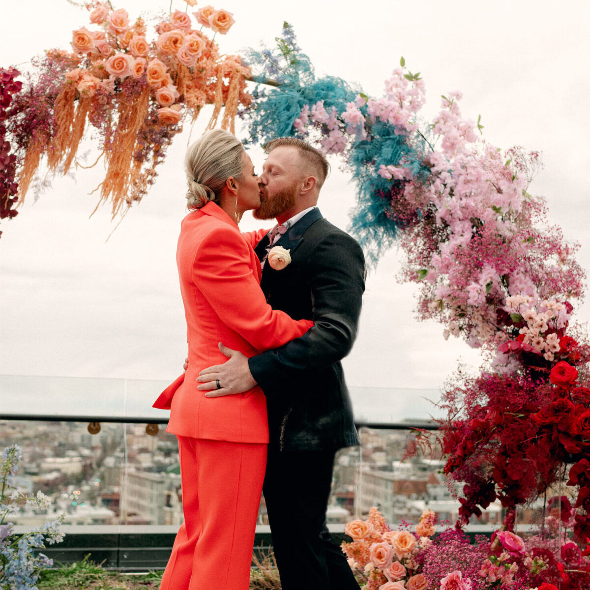 Wedding couple kissing under their vibrant floral arch at their colorful surprise rooftop wedding.