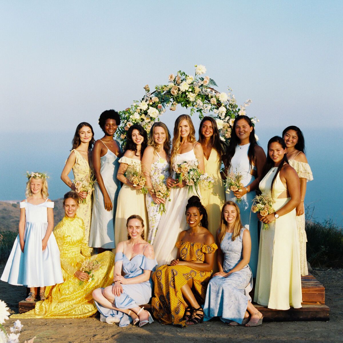 Mismatched bridesmaid dresses: Bride with bridesmaids in mismatched bridesmaid dresses standing in the sunlight in front of a floral wedding arch