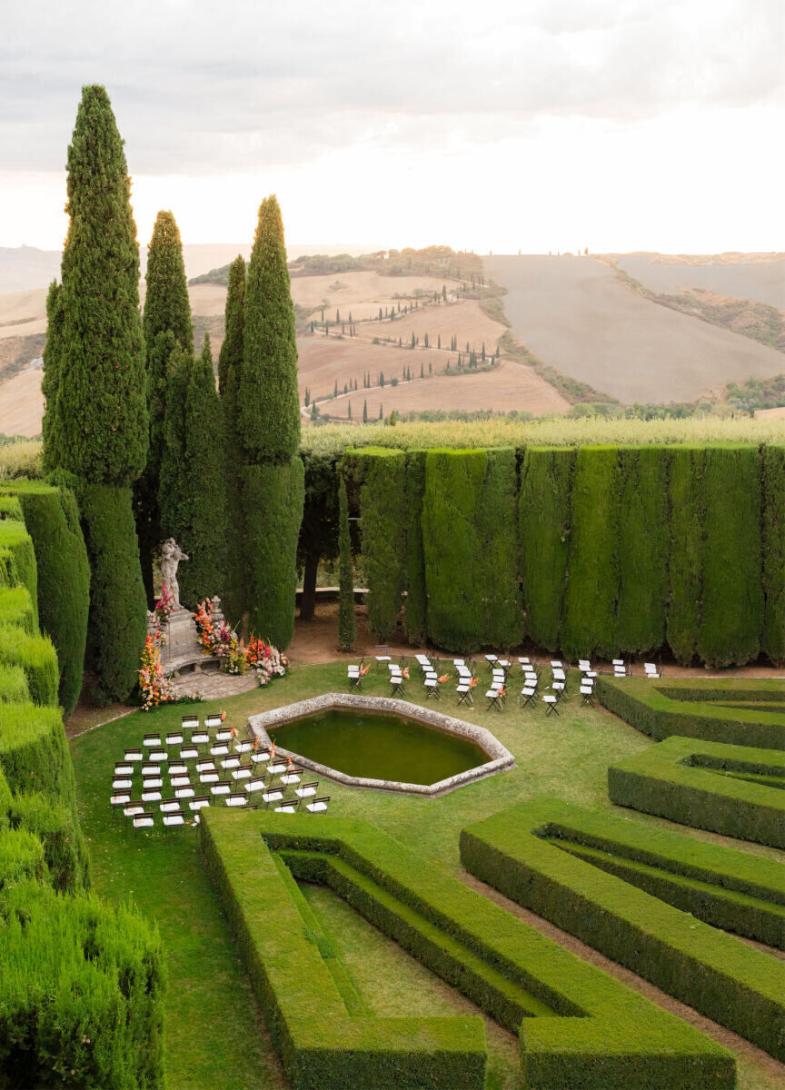 A Tuscan wedding ceremony in an Italian garden with chairs set facing a floral-covered statue.