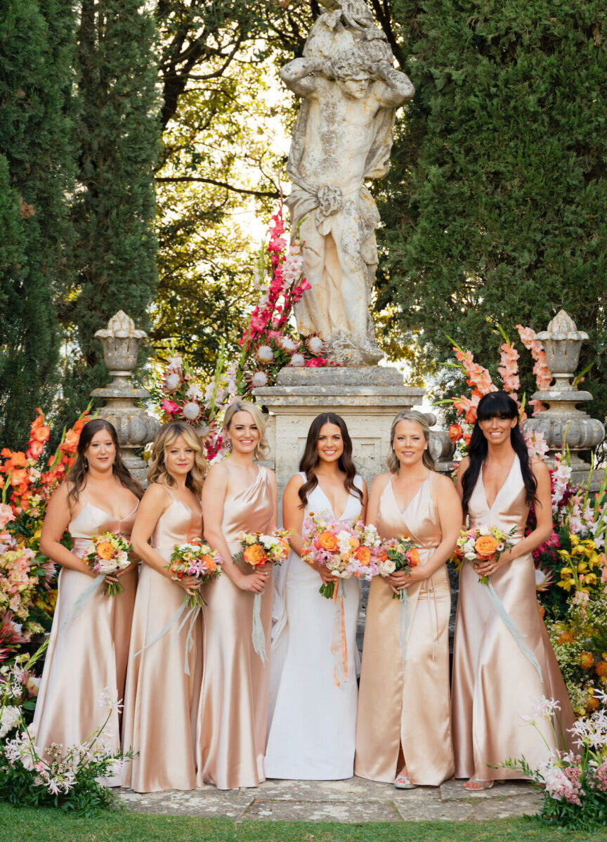 A bride surrounded by her bridesmaids (wearing satin peach dresses) hold vibrant bouquets and pose for a portrait in the garden where the Tuscan wedding ceremony took place.