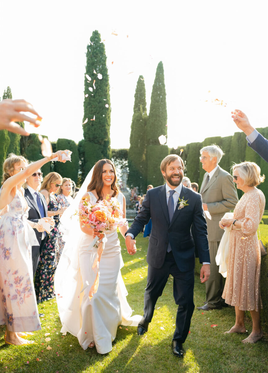 A couple smiles as they recess up the aisle under a shower of flower petals at their Tuscan wedding ceremony in a Cypress tree-lined garden.