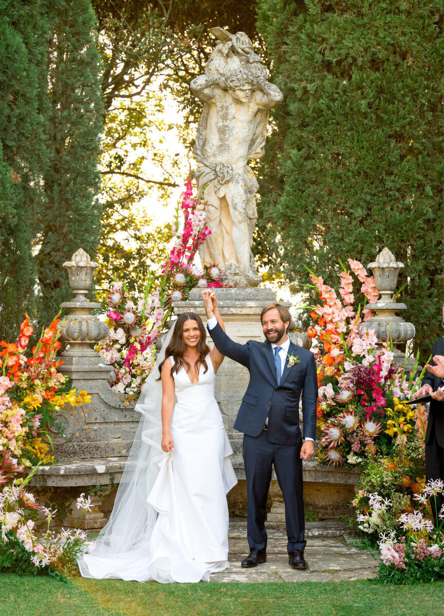 A bride and groom clasp hands and raise them in excitement at the conclusion of their Tuscan wedding ceremony, which took place in a garden decorated with colorful flowers around a statue.