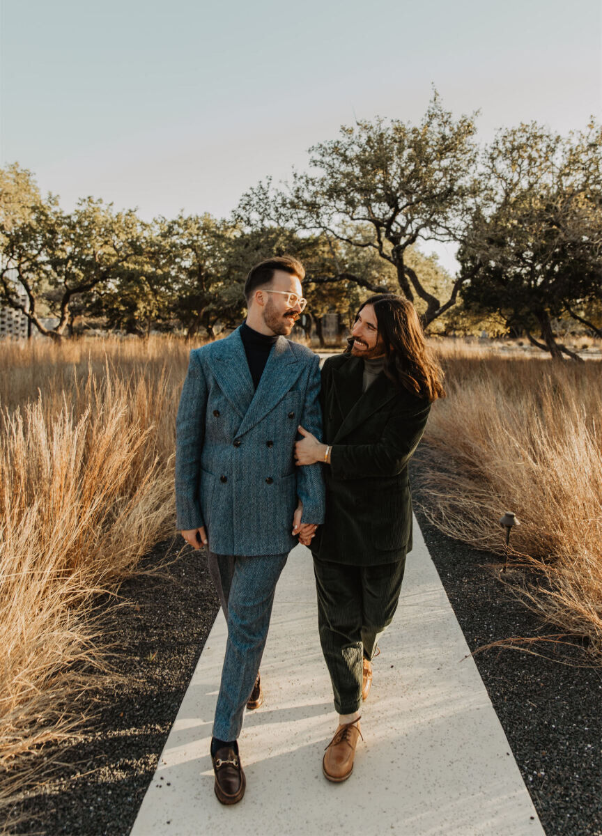 A pair of grooms strolls down the pathway of their Texas wedding venue holding hands and smiling widely.